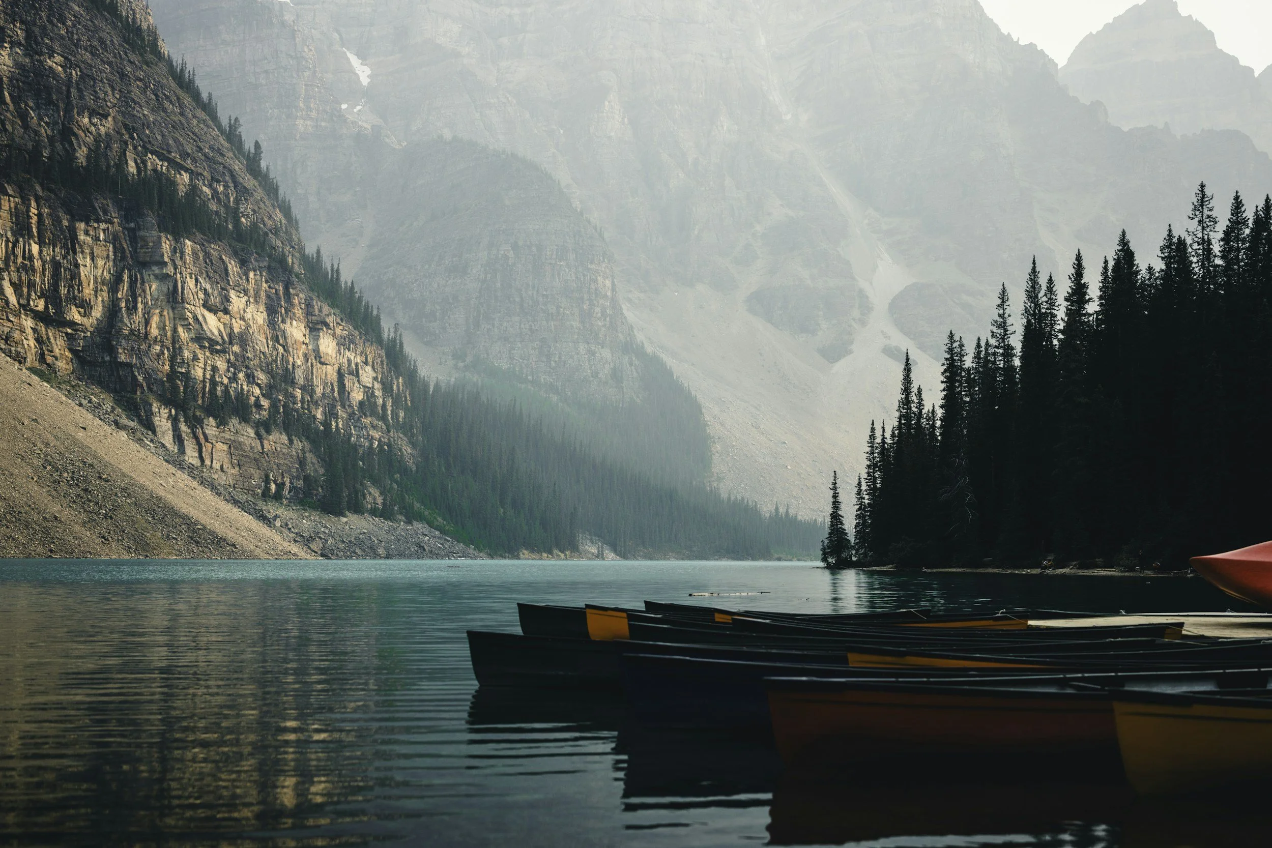 A serene lake surrounded by towering mountains with snow patches, dense evergreen trees, and kayaks floating on the water.