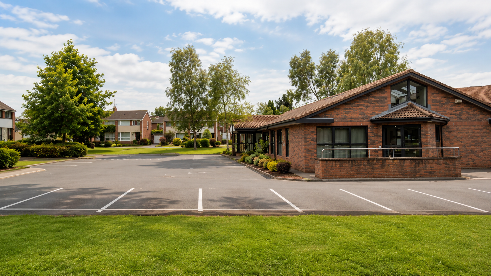 A parking lot outside a residential building with well-maintained gardens and trees, under a partly cloudy sky.