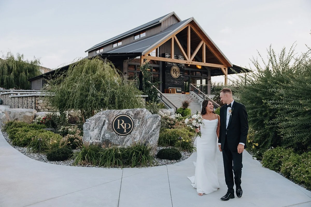 A newlywed couple walks happily outside a modern house with wooden accents. The bride wears a white wedding dress and holds a bouquet, while the groom wears a black tuxedo. They are surrounded by greenery and colorful flowers.