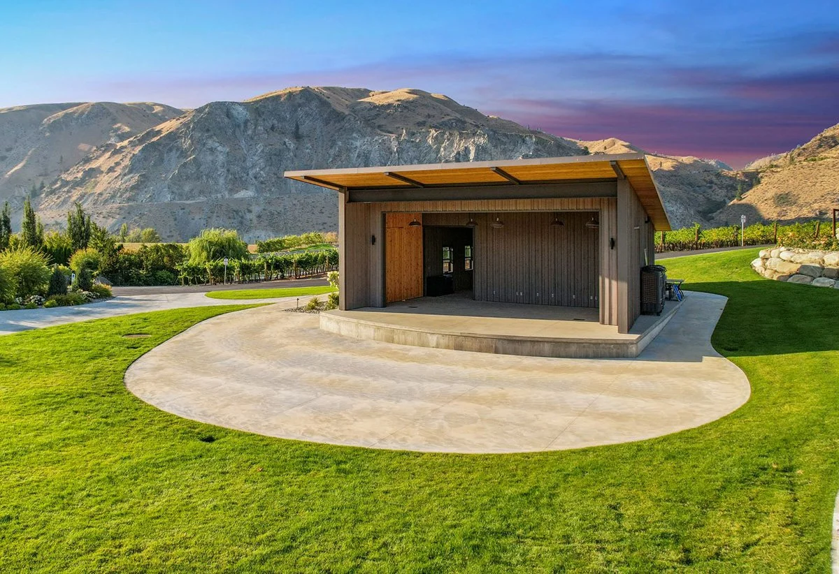 An outdoor stage with a wooden roof in a lush green park, mountains in the background, evening sky with purple and blue hues.