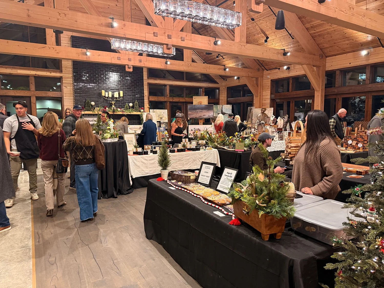 Indoor holiday market with various vendor tables selling arts, crafts, and holiday decorations. People browsing and shopping in a wooden, spacious room with a vaulted ceiling and large windows.