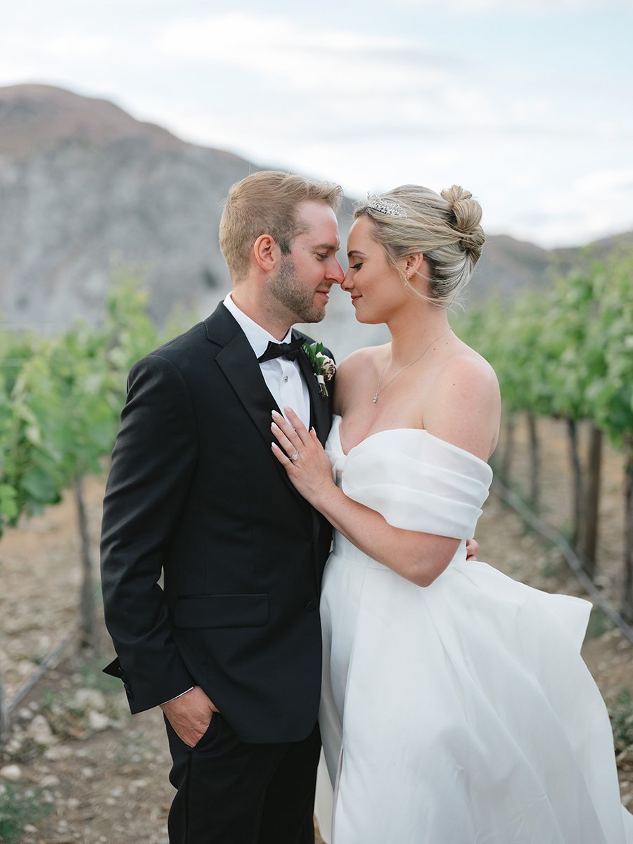 A bride and groom standing close together in a vineyard, about to touch foreheads, with mountains in the background.