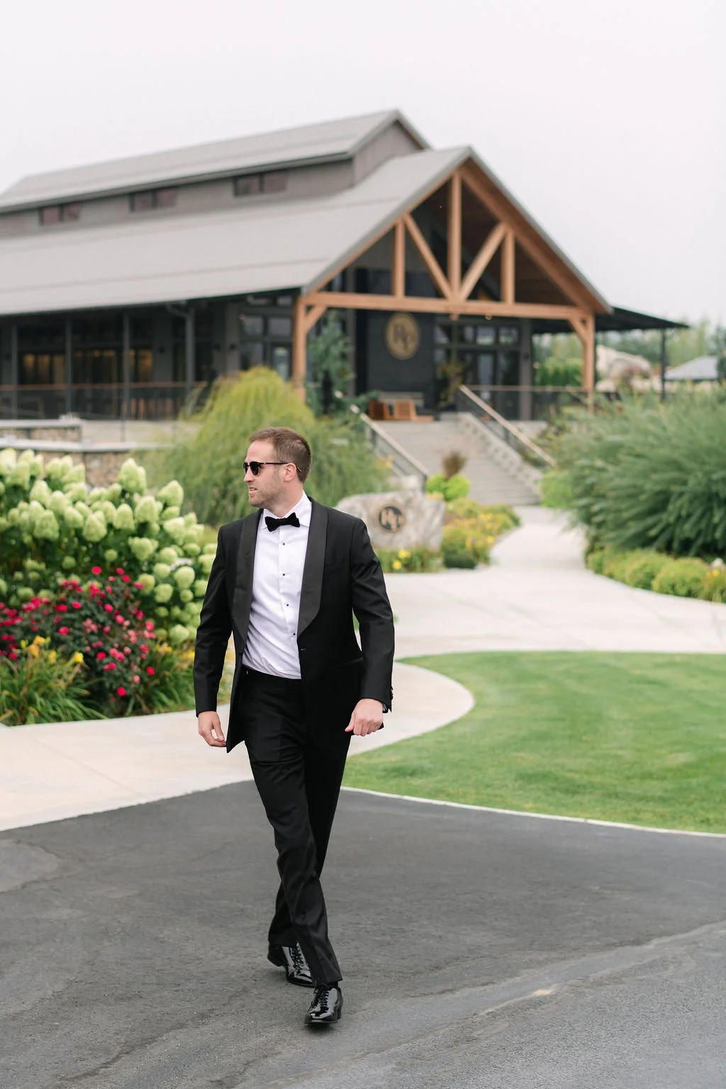 Man in tuxedo and sunglasses walking outdoors near a modern house with landscaped garden.