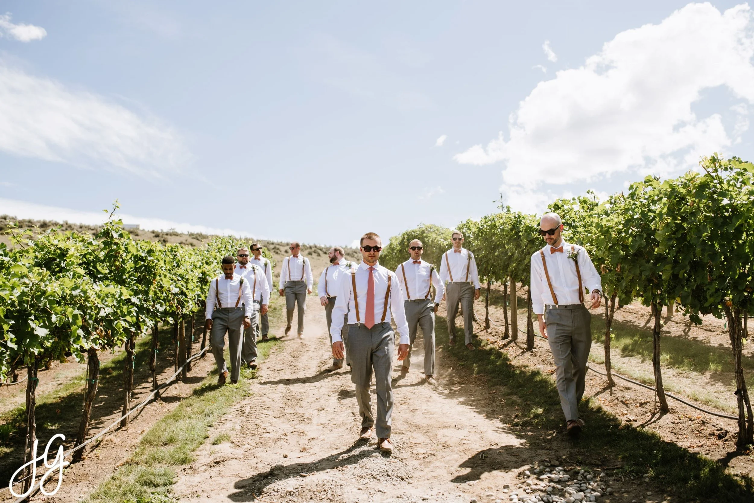 Group of men dressed in formal attire walking through a vineyard under a partly cloudy sky.