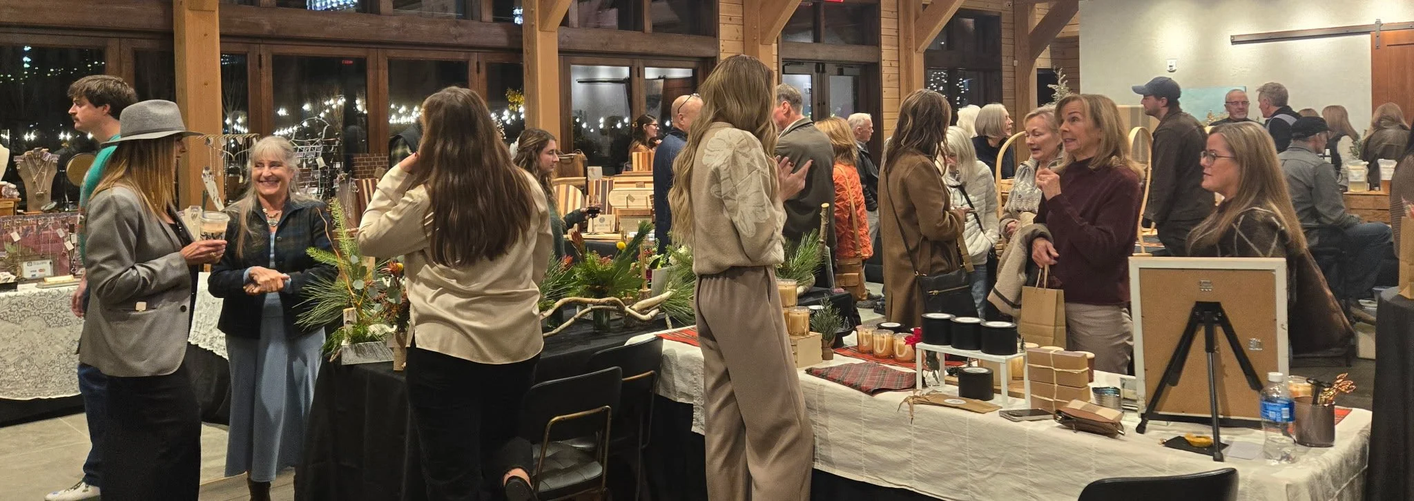 People socializing and shopping at an indoor market or crafts fair in a warmly lit wooden building, with tables displaying various handmade items and decorations.