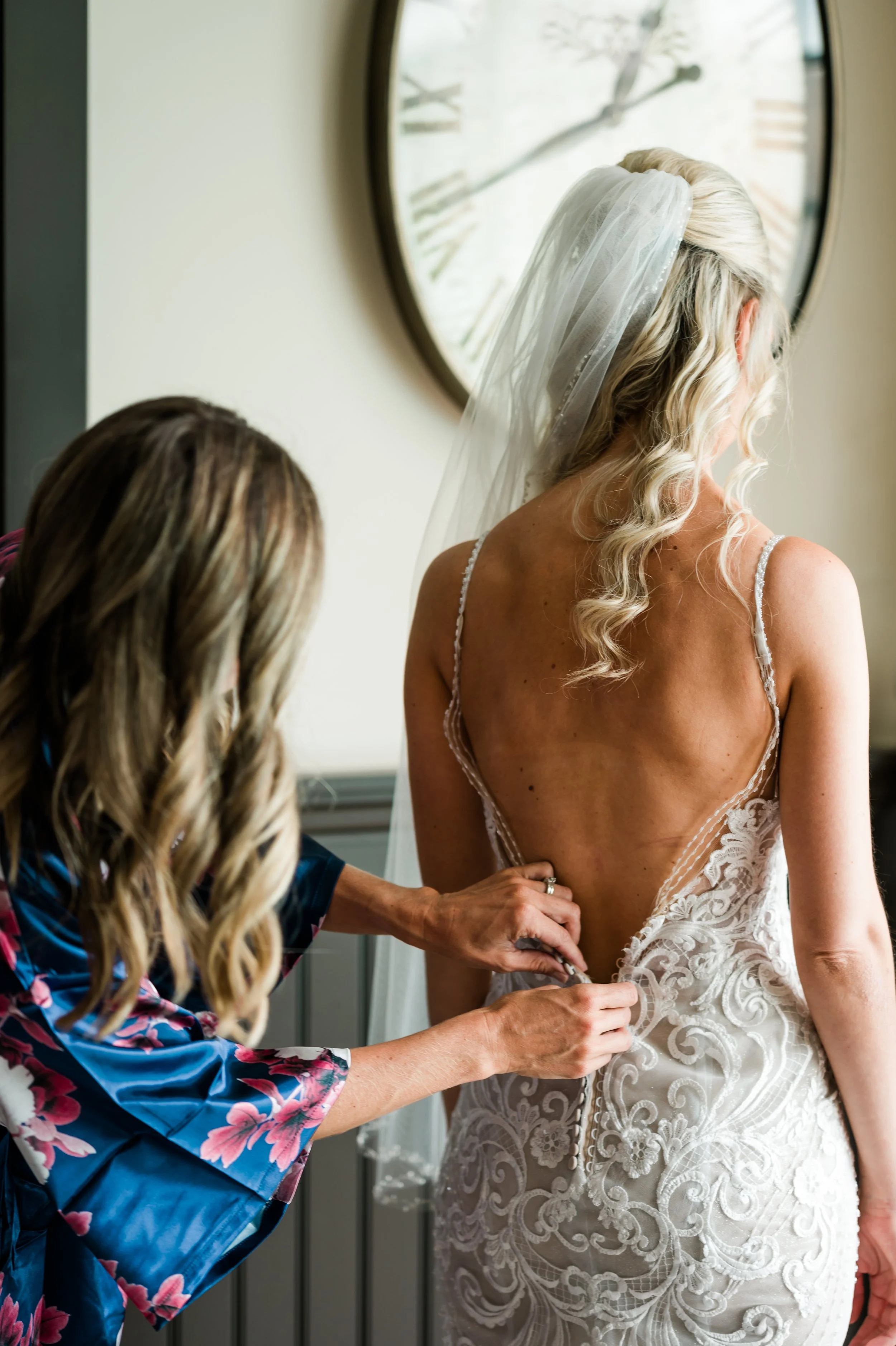 A woman in a wedding dress has her dress buttoned up as another woman helps her. A large round mirror reflects a window with trees outside.