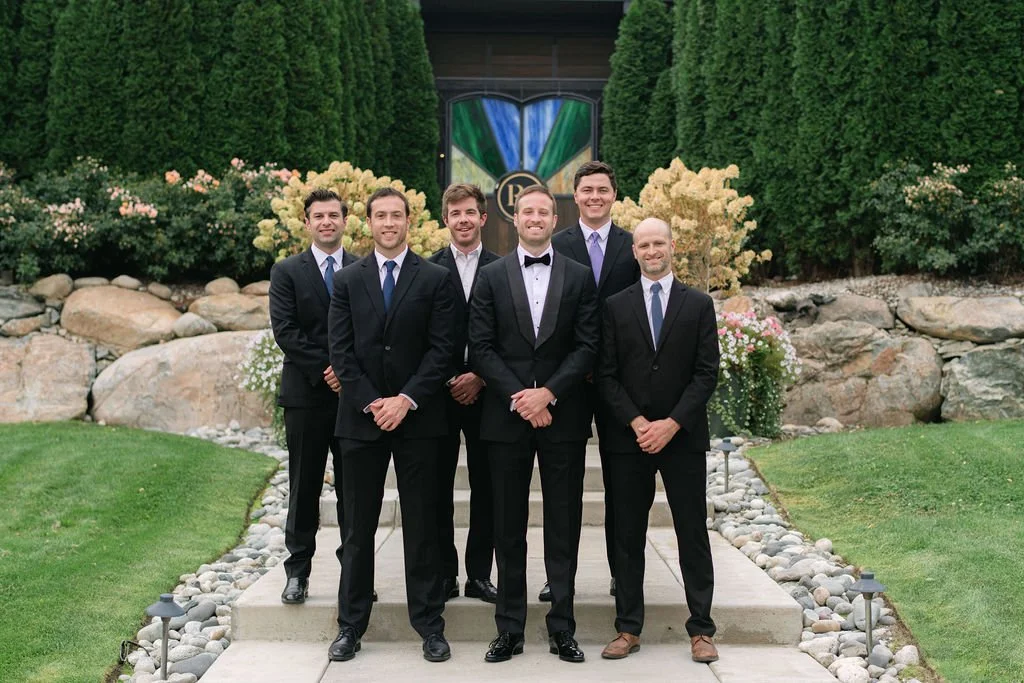 Six men dressed in suits and tuxedos standing outdoors on steps, smiling, with a garden and stained glass window in the background.
