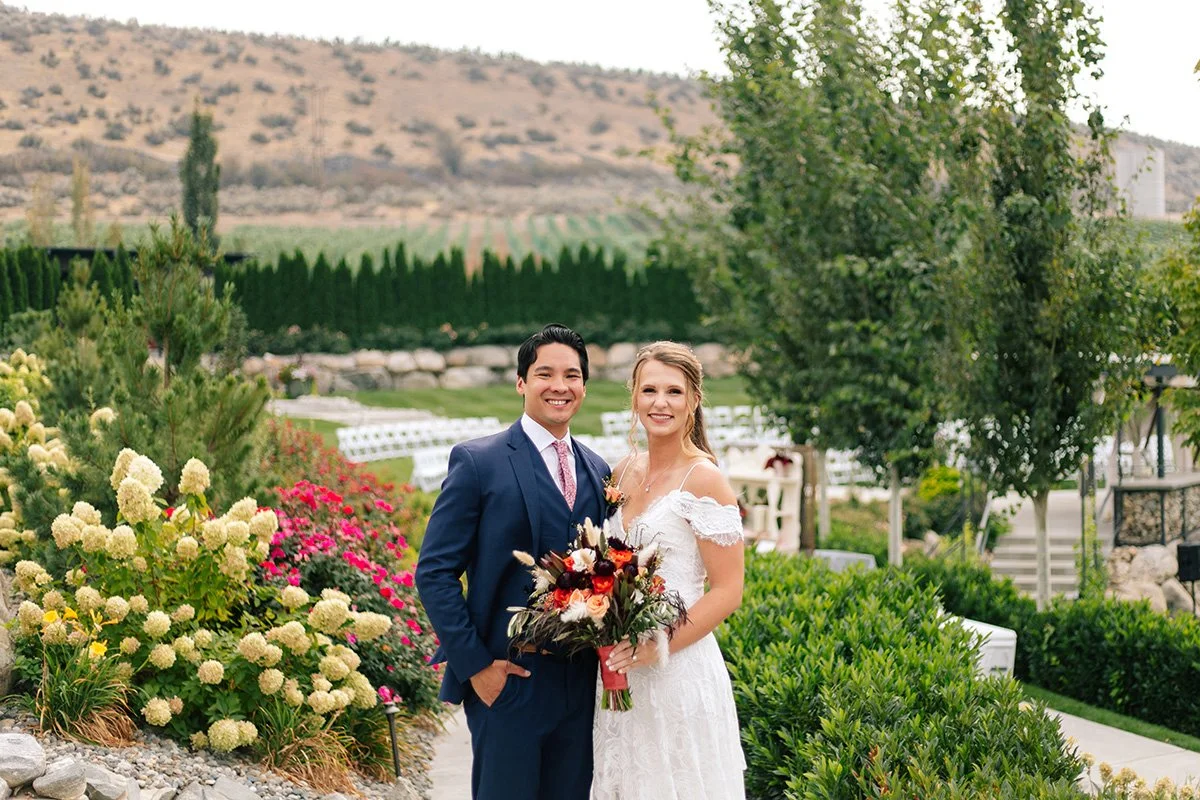 A smiling couple, the bride in a white wedding dress holding a bouquet and the groom in a dark suit, standing in a lush garden with colorful flowers and greenery.