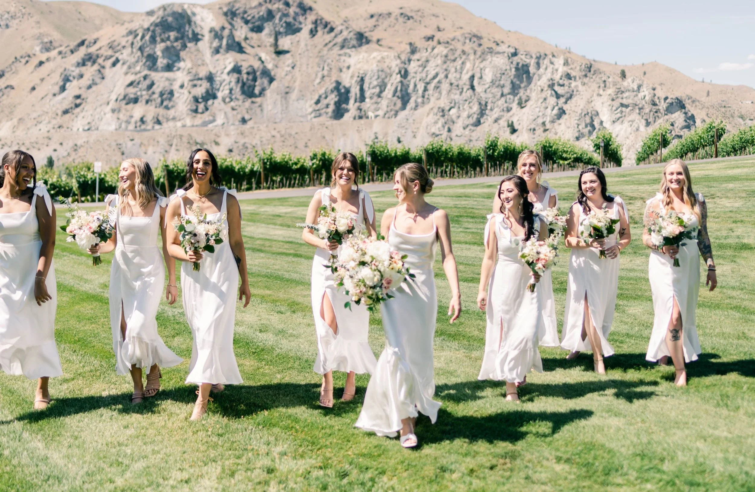 A group of women in white dresses walking on grass, holding bouquets, with a backdrop of mountains and a vineyard.