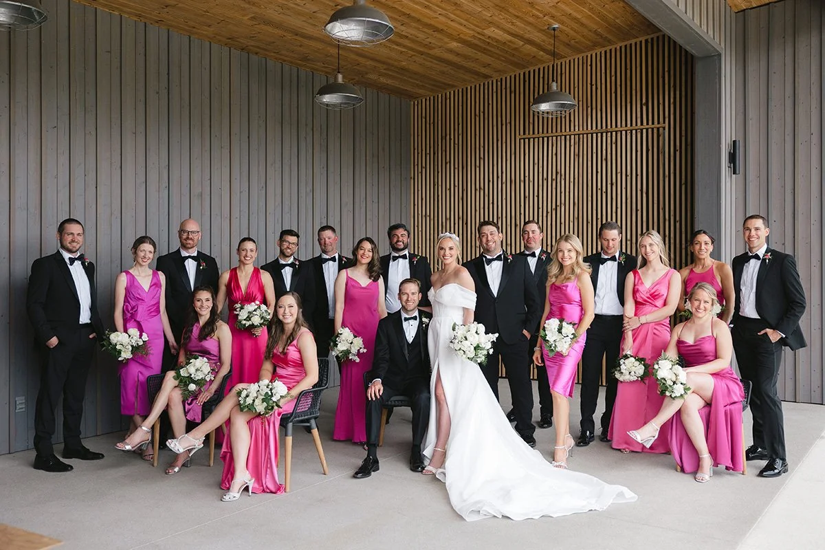 A group of people dressed in formal wedding attire, including a bride in a white wedding gown and bridesmaids in pink dresses, posing for a photo indoors against wooden wall panels.