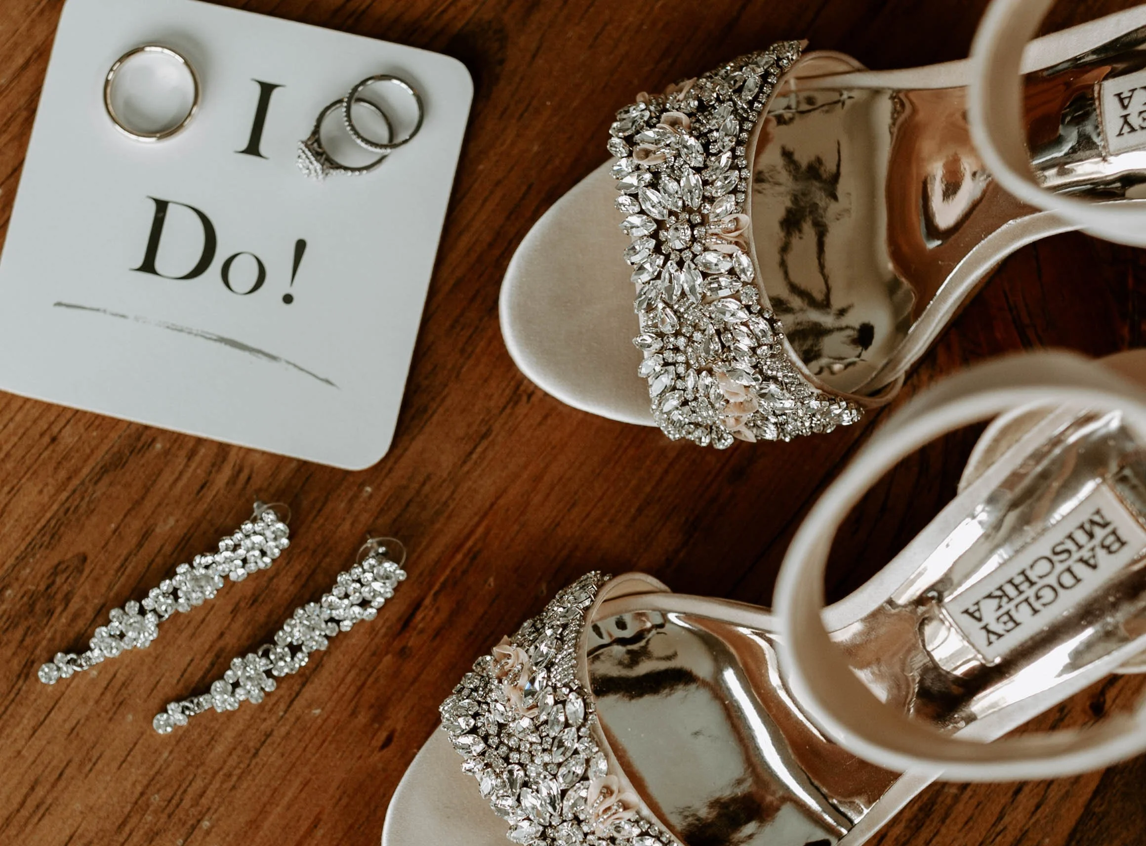 Close-up of jewelry including a sparkling bracelet, earrings, and additional jewelry pieces on a wooden surface, with a small card that reads "I Do!" positioned nearby.