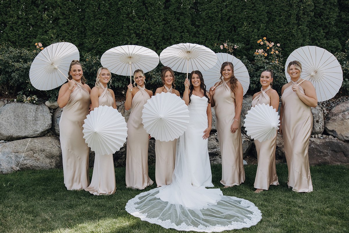 Group of eight women at a wedding, dressed in beige gowns, holding white parasols, standing on grass in front of rocks and greenery, with the bride in the center wearing a white gown and veil.
