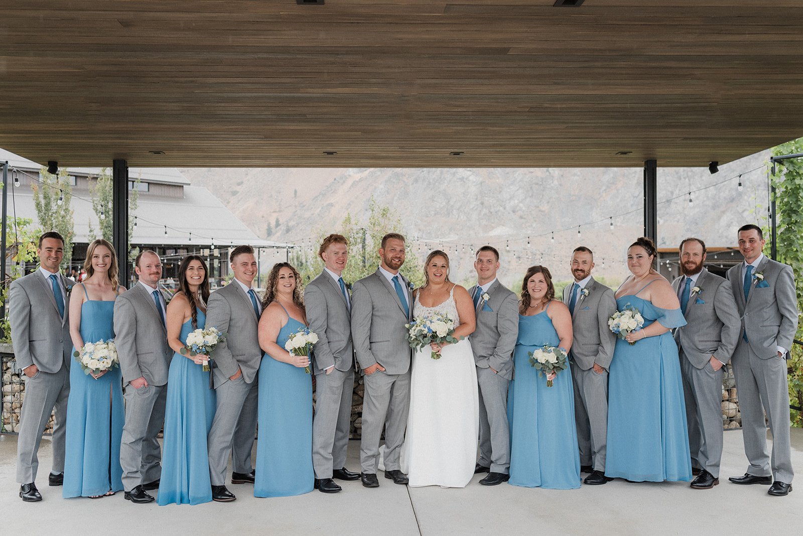 Group of 15 people at a wedding, dressed in formal attire, standing outdoors under a wooden ceiling. The women are wearing light blue dresses and holding white bouquets, while the men are wearing light gray suits with blue ties. The bride is in a whi