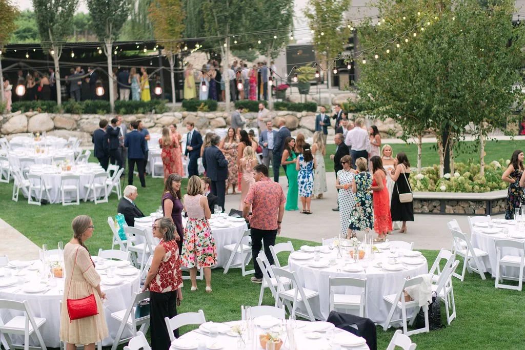 Outdoor wedding or formal event with people mingling, white tables and chairs, string lights, and a garden setting.