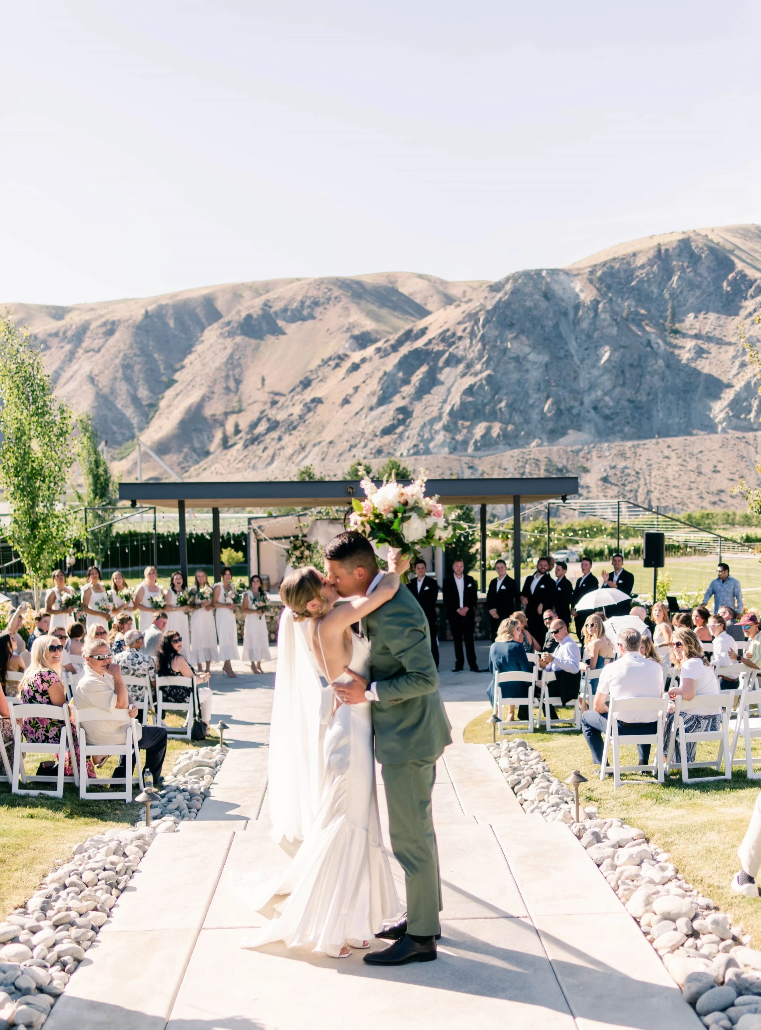 A bride and groom kiss at an outdoor wedding ceremony with mountains in the background. Guests are seated and standing, watching the couple. The bride is holding a bouquet of flowers, and the groom is wearing a suit. Bridesmaids and groomsmen are lin