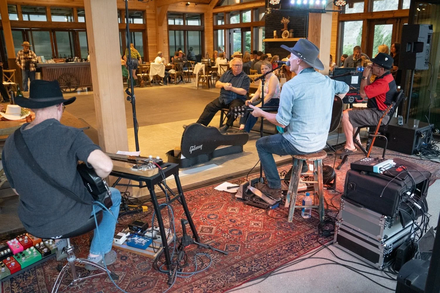 A live band performing indoors at a rustic wooden venue, with multiple musicians playing guitars and other instruments, and an audience seated in the background.