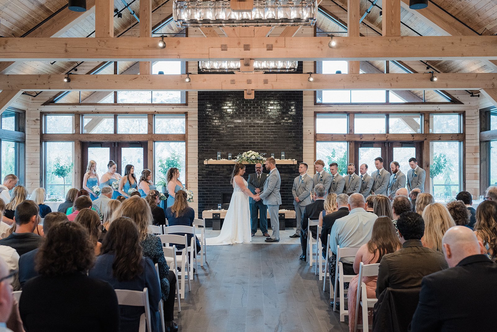 A wedding ceremony taking place in a modern wooden chapel with large windows. The bride and groom stand at the altar holding hands, facing each other, with the officiant between them. Bridesmaids in blue dresses stand to the left, and groomsmen in gr