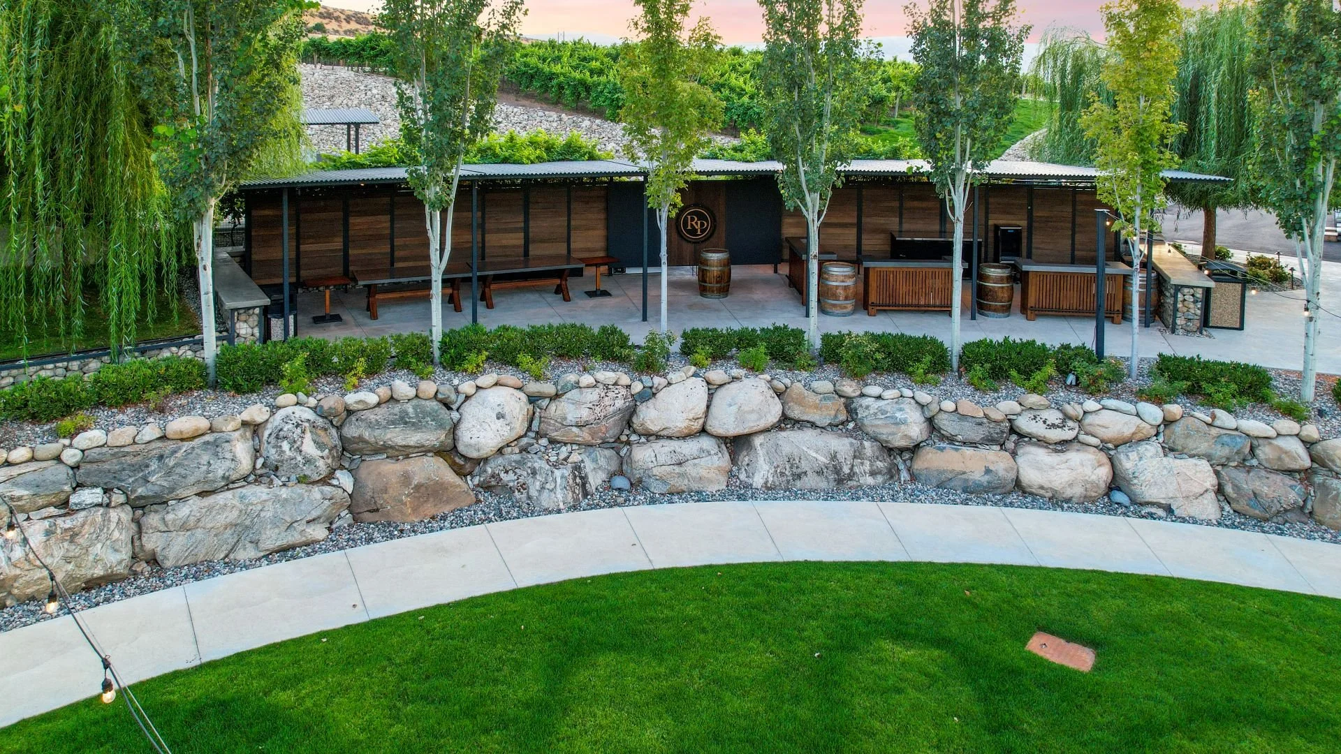 Outdoor seating area with benches, barrels, and a grill, surrounded by trees, rocks, and greenery.