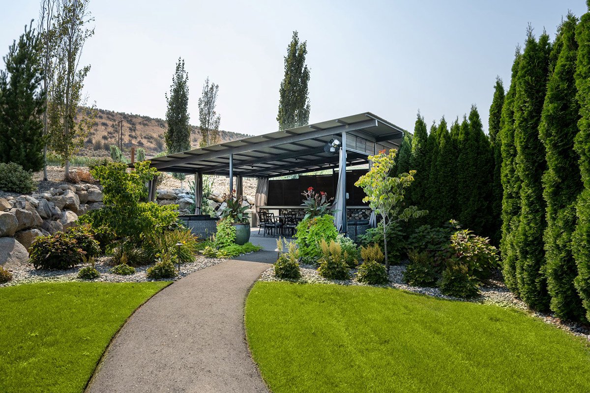 A backyard garden with a pathway leading to a covered patio, surrounded by trees and shrubs.
