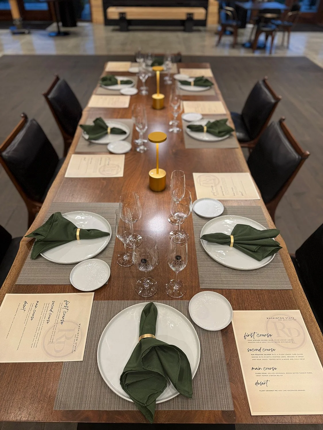 A restaurant table set for a meal, with white plates, green napkins tied with gold bands, wine glasses, small plates, and printed menus on beige placemats.