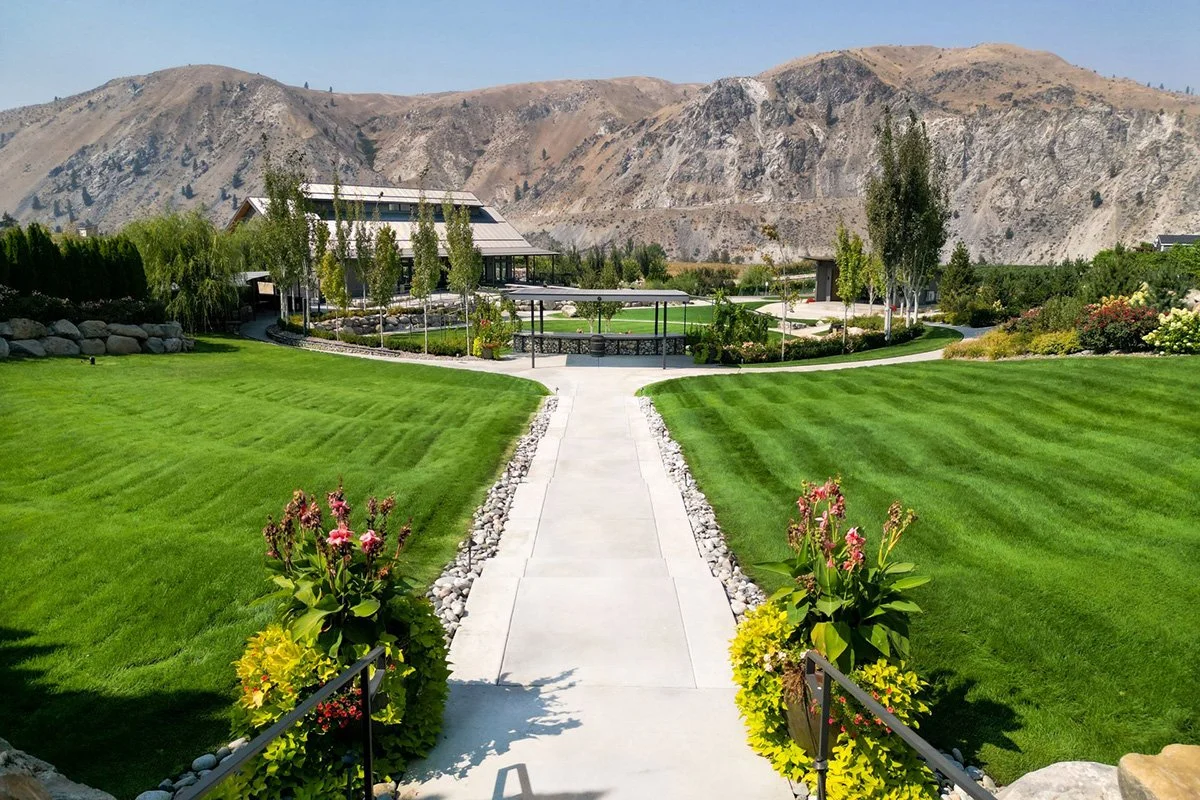 A lush green lawn with a central walkway, flowering plants in pots, trees, and a building with mountains in the background.