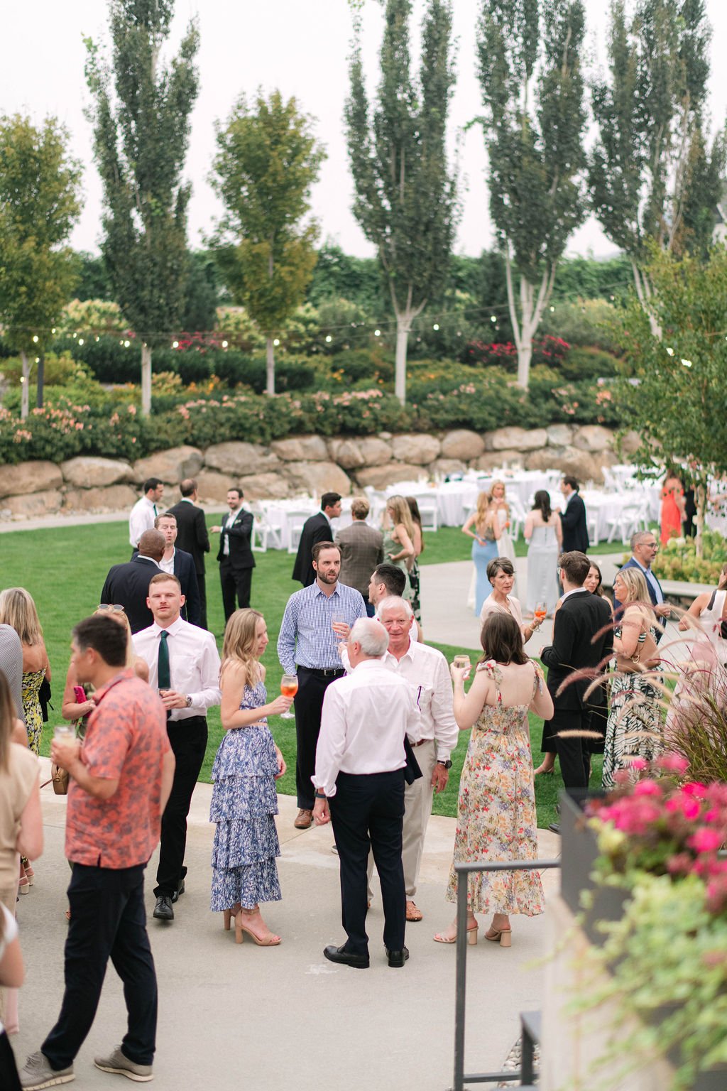 Group of people socializing outdoors at a garden party or wedding reception, with tables set for dining, trees, and string lights in the background.