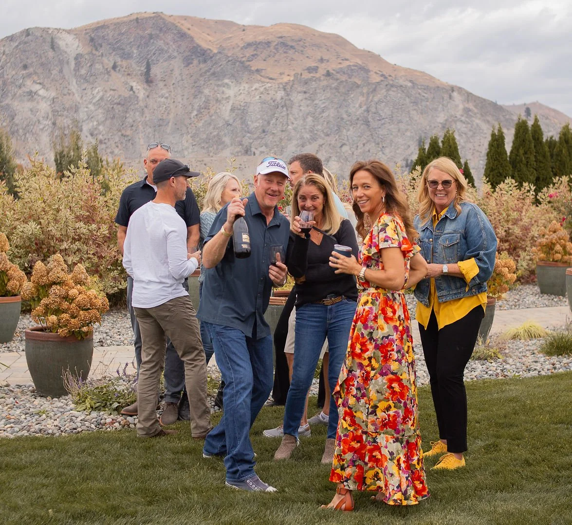 Group of people celebrating outdoors with drinks in front of mountains and potted plants on a lawn.