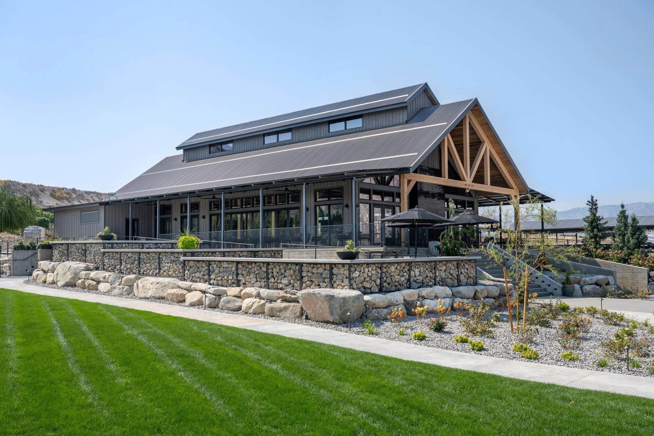 Modern house with metal roof, large windows, and wooden architectural details, surrounded by landscaped garden, rocks, and a green lawn under a clear blue sky.