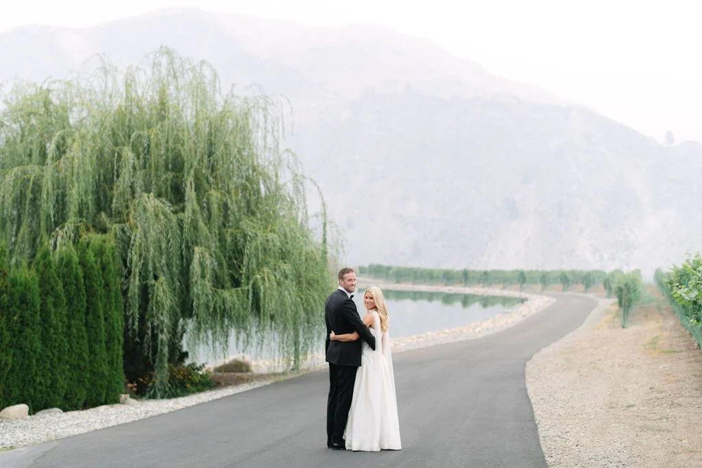 A bride and groom stand together on a paved path beside a body of water, with green trees and mountains in the background.