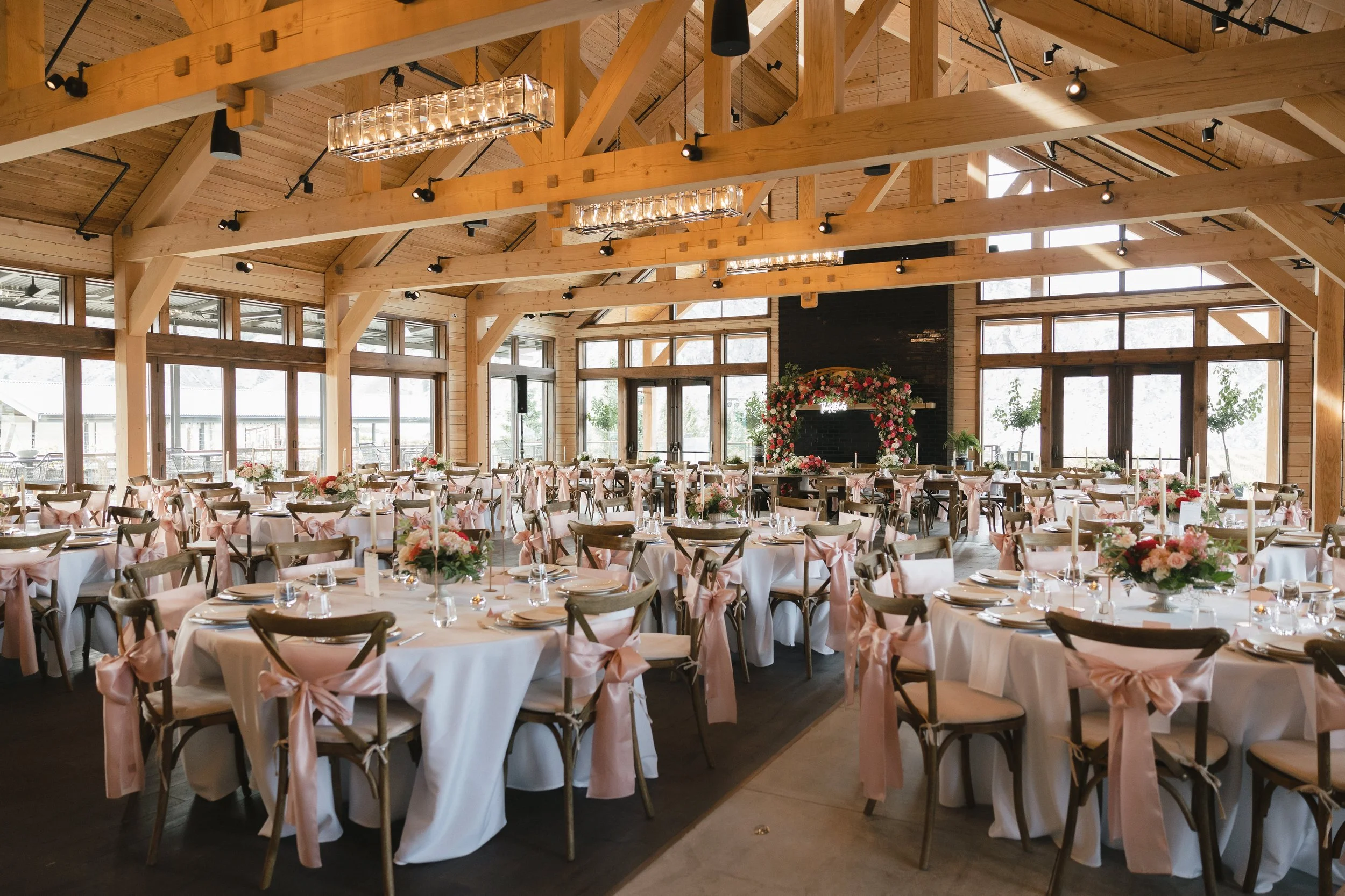 Elegant wedding reception setup with round tables covered in white linens, decorated with pink bows and floral centerpieces, in a spacious wooden venue with large windows and modern light fixtures.