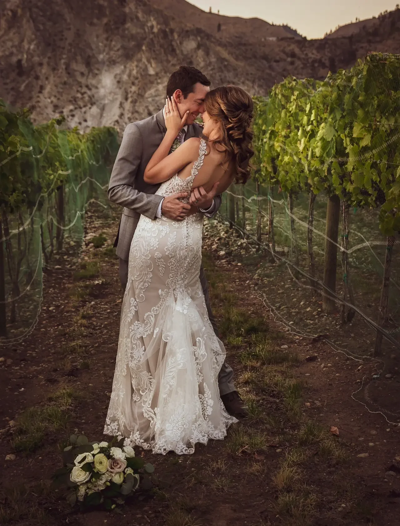 A happy couple in wedding attire embracing in a vineyard during sunset, with a bouquet of flowers on the ground nearby.