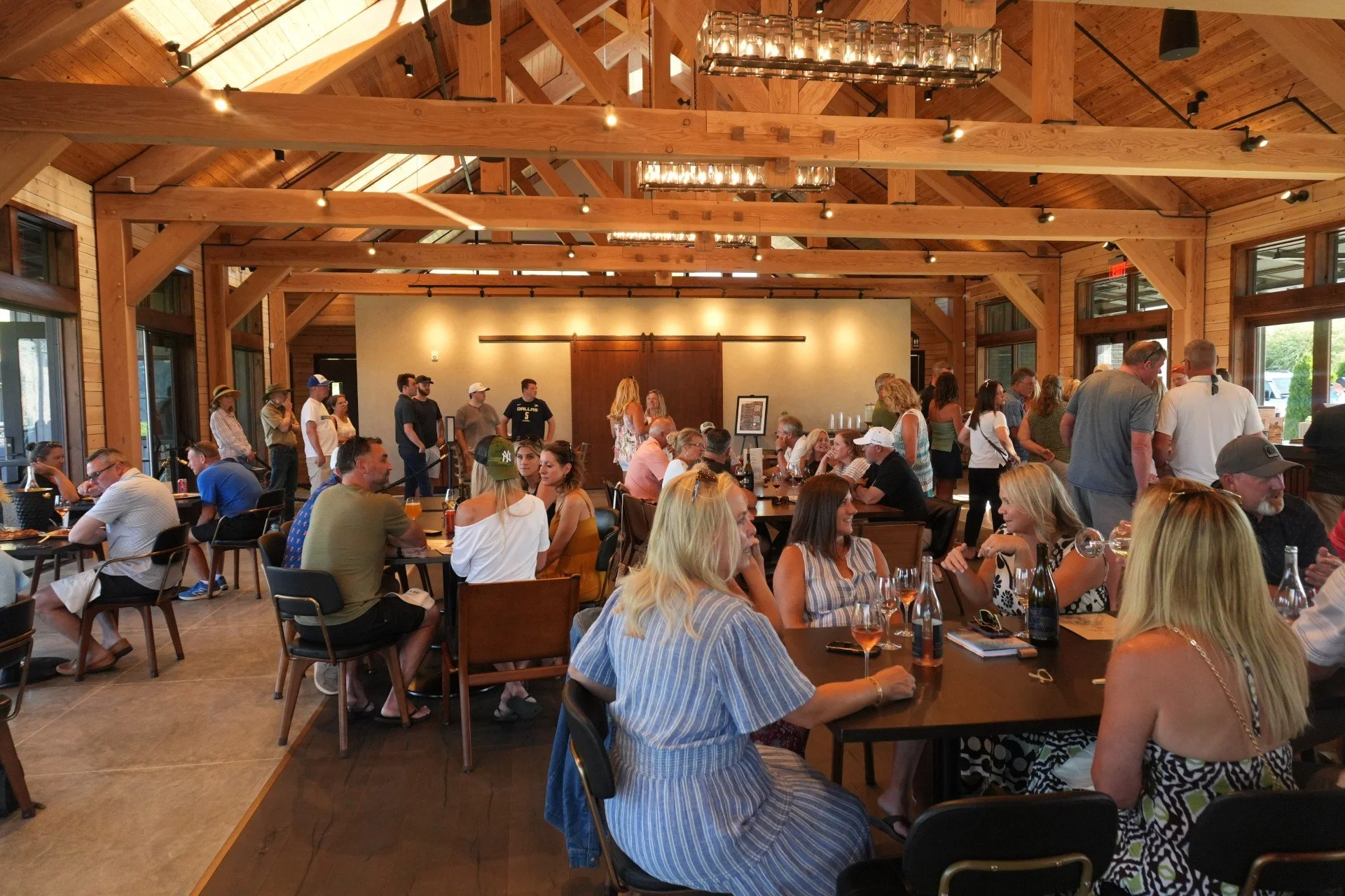 People socializing and dining in a spacious, rustic-style restaurant with wood-paneled walls and ceiling, large windows, and modern lighting fixtures.