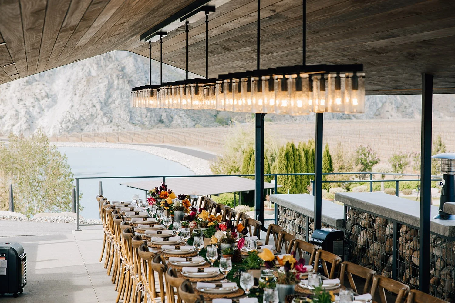 A long banquet table set for a meal, decorated with colorful flowers and neatly arranged plates, glasses, and utensils, on a covered outdoor patio with scenic mountain and vineyard views.