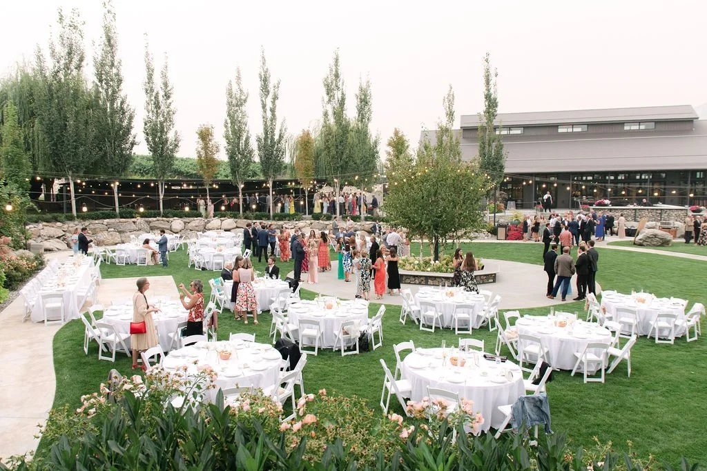 Outdoor wedding reception with round tables covered in white cloths and white chairs, people socializing, string lights overhead, trees, and a modern building in the background.