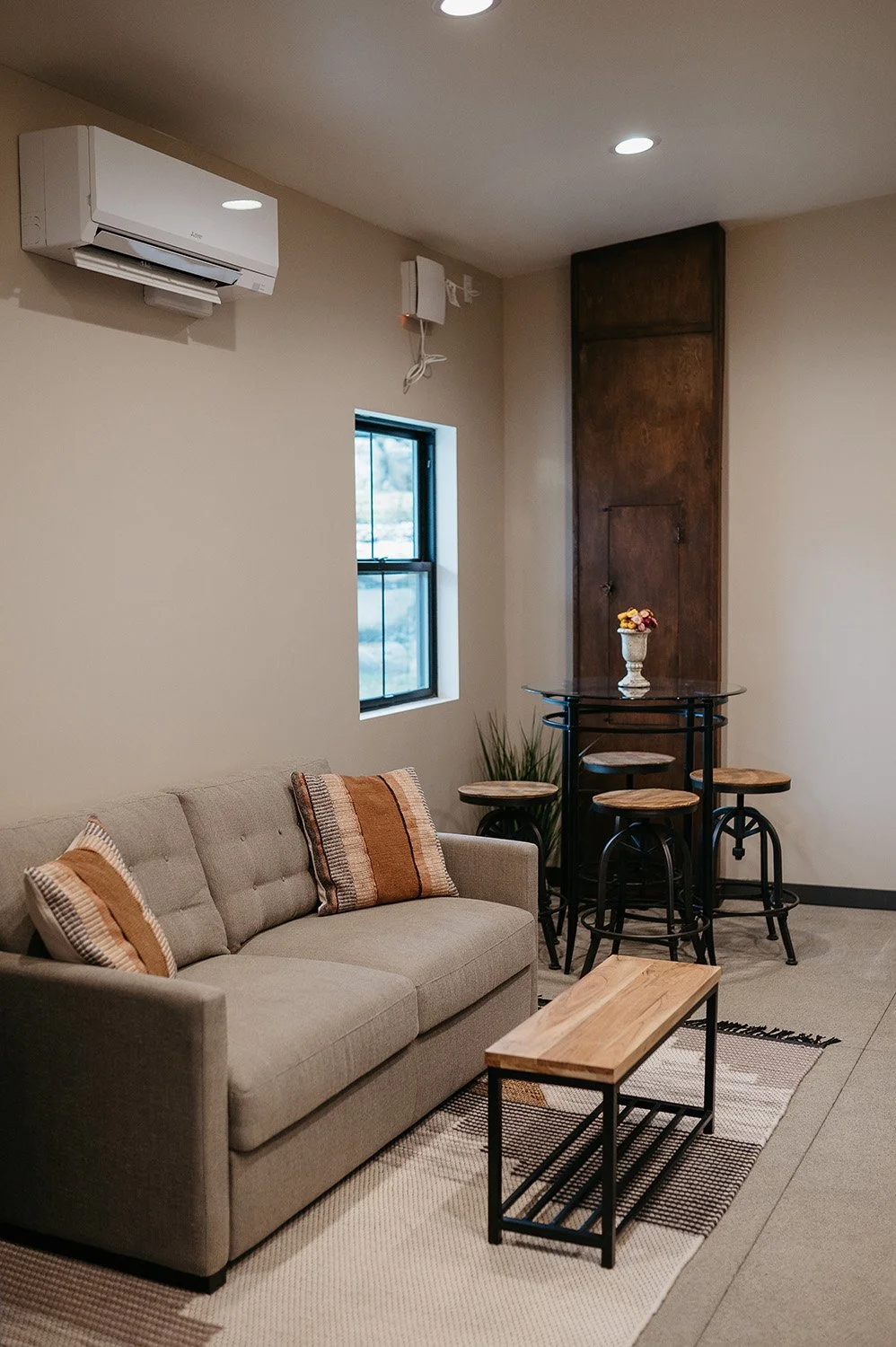 Living room with beige sofa, two striped pillows, a small wooden coffee table, a round high-top table with four stools, a small window, a flower vase on the table, a potted plant, a wooden panel wall, an air conditioning unit, and round ceiling lights.
