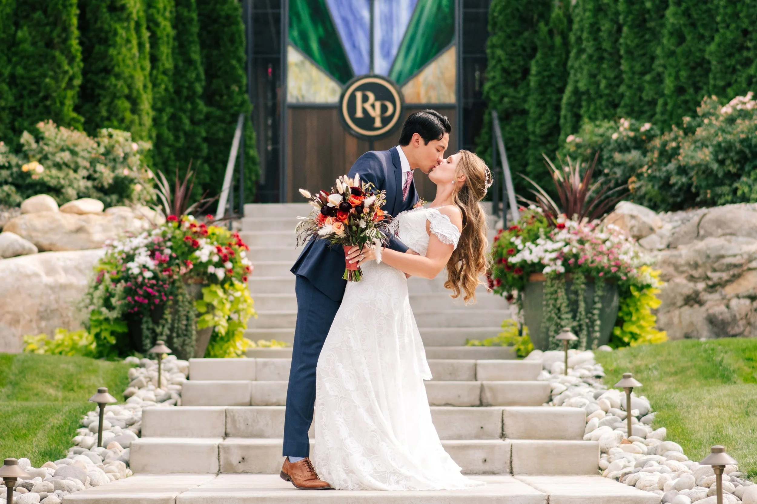 A couple in wedding attire sharing a kiss on outdoor steps surrounded by flowers and greenery.