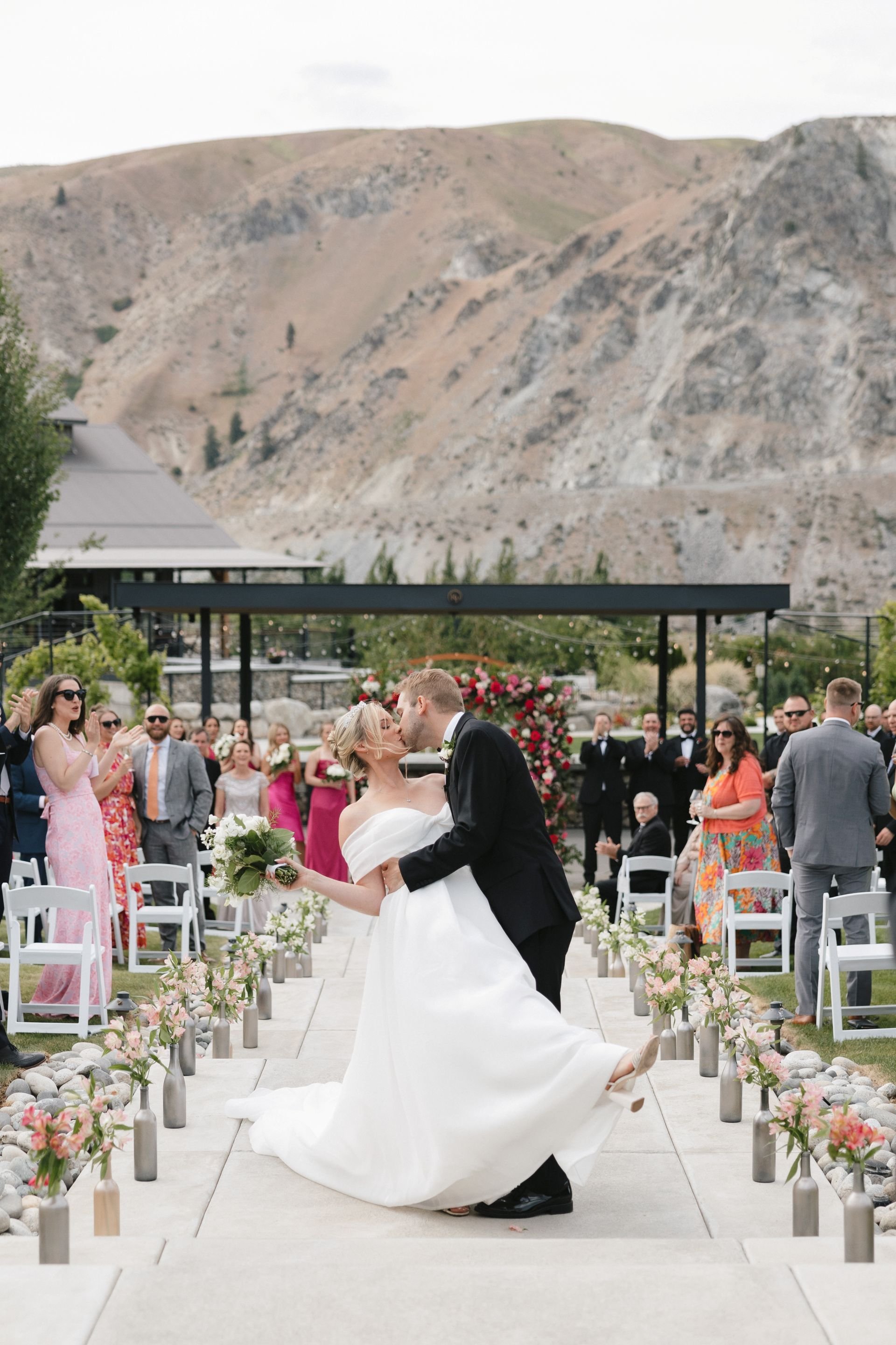 A couple gets married outdoors with a mountain backdrop. The bride wears a white wedding gown and the groom in a black suit. They are kissing at the altar, surrounded by their guests who are clapping and smiling.
