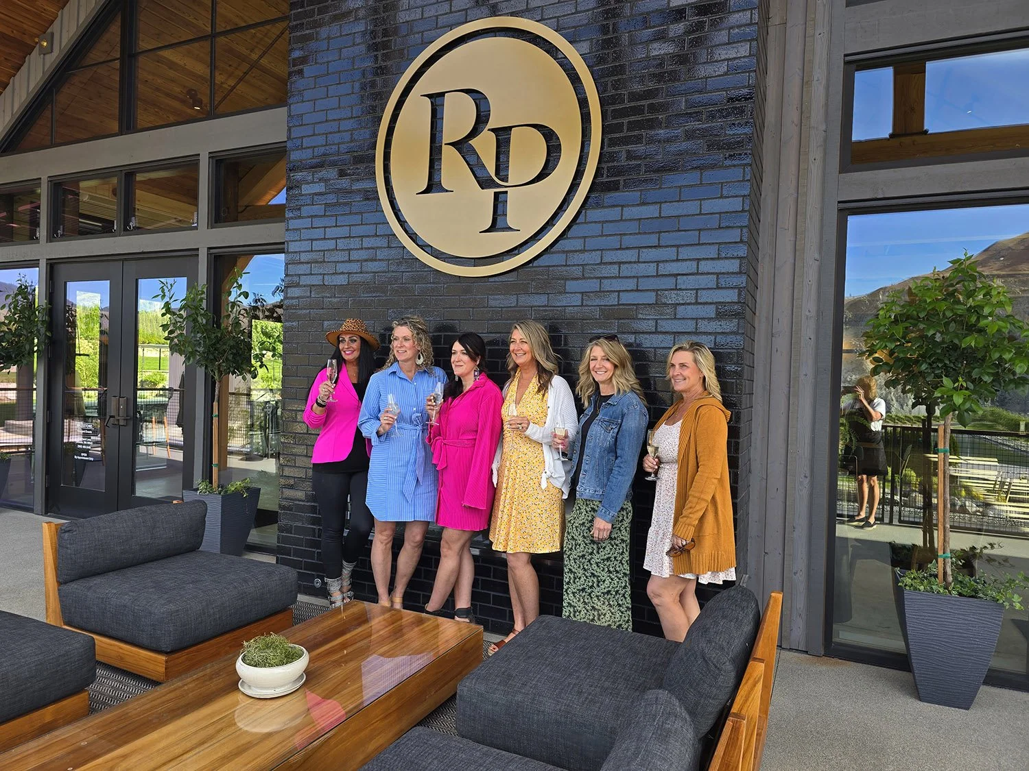 Six women standing together indoors, smiling, holding glasses of wine, with large RD logo on the dark brick wall behind them.