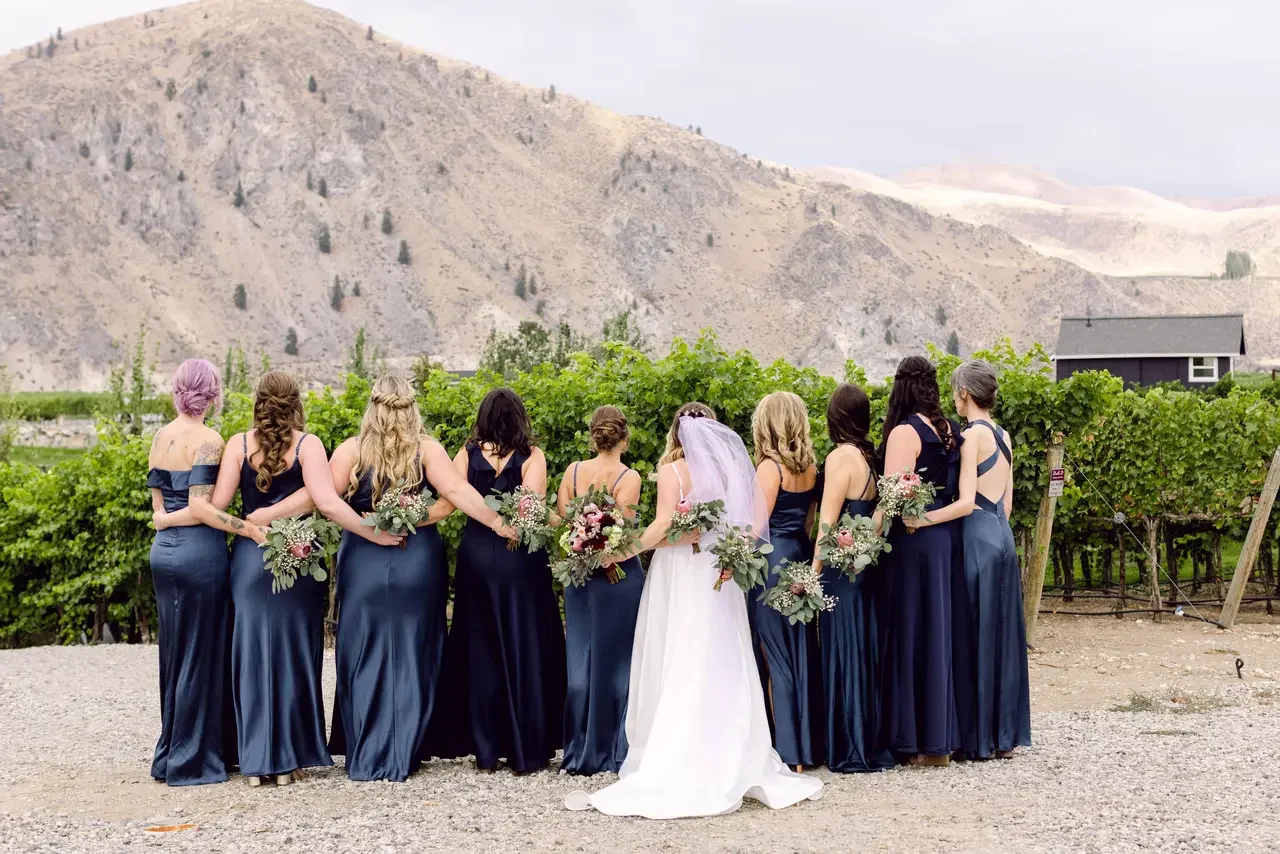 A bride in a white wedding gown with a veil standing among bridesmaids in navy blue dresses, holding bouquets, in a vineyard with mountains in the background.