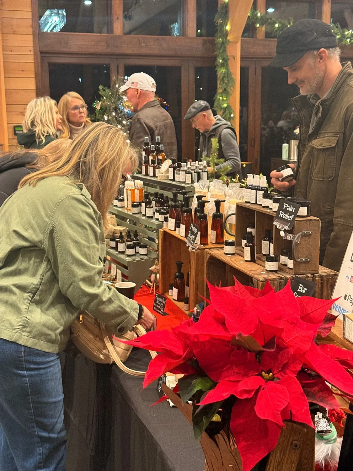 People shopping at a holiday gift market with wellness products, including essential oils and natural remedies, on a table decorated with poinsettias, inside a warmly lit wooden venue.