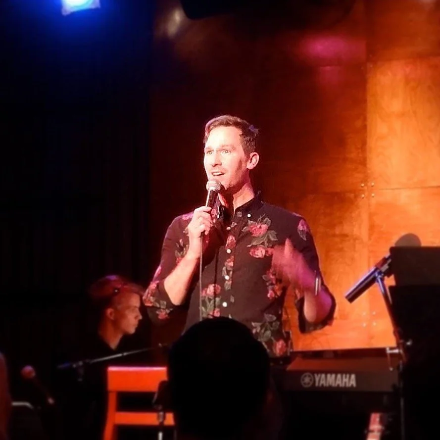 A man singing into a microphone on stage with a keyboard and a woman playing piano in the background, illuminated by stage lighting.