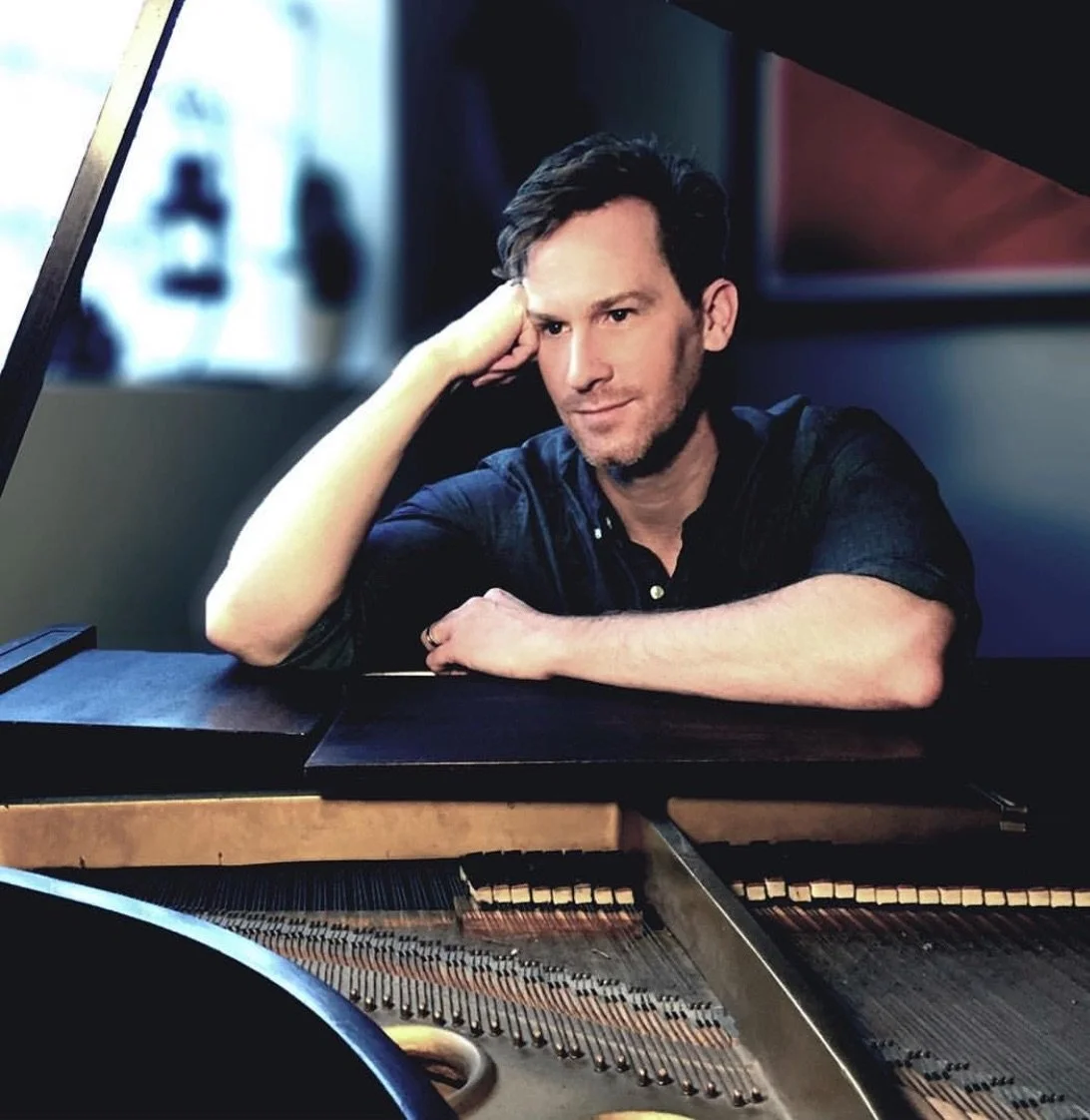 A man with dark hair and light stubble, resting his head on his hand, sitting beside an open grand piano.
