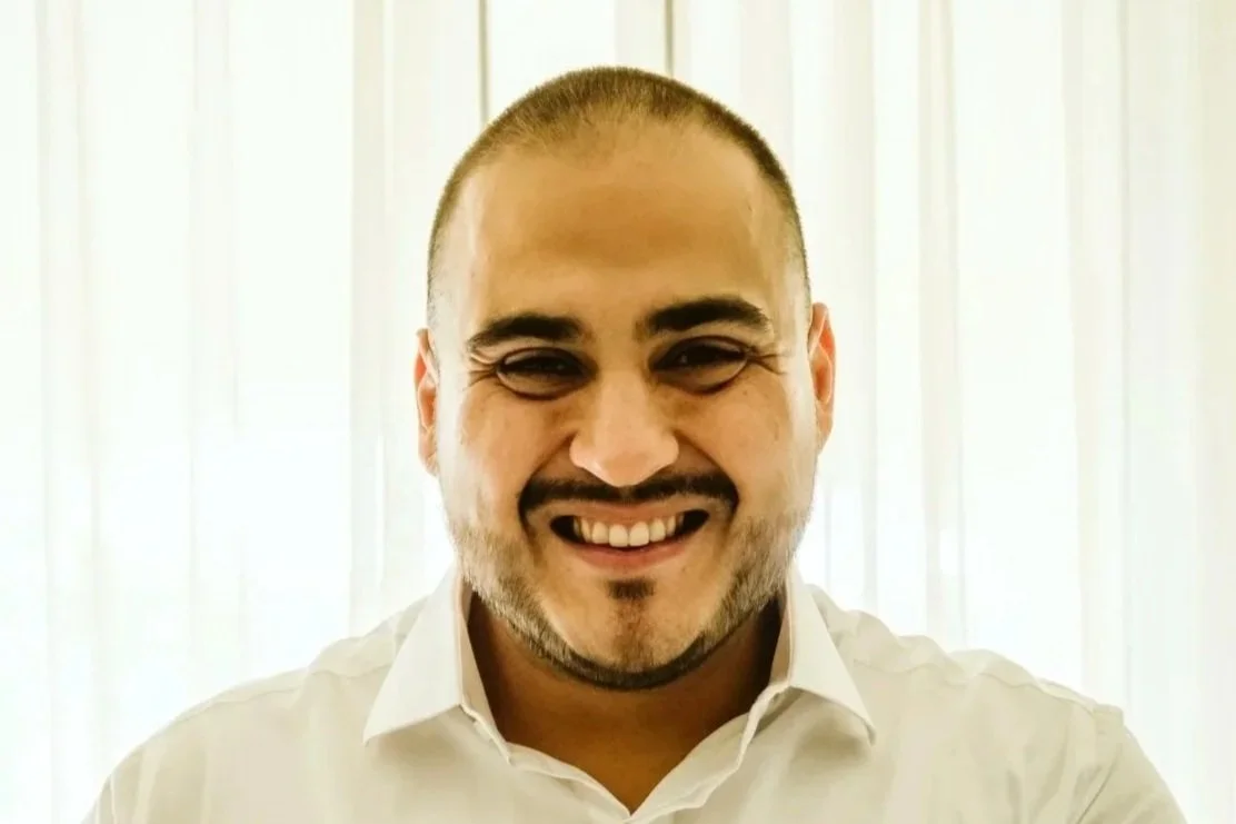 Smiling man with short hair, beard, and white shirt inside a room with white curtains.