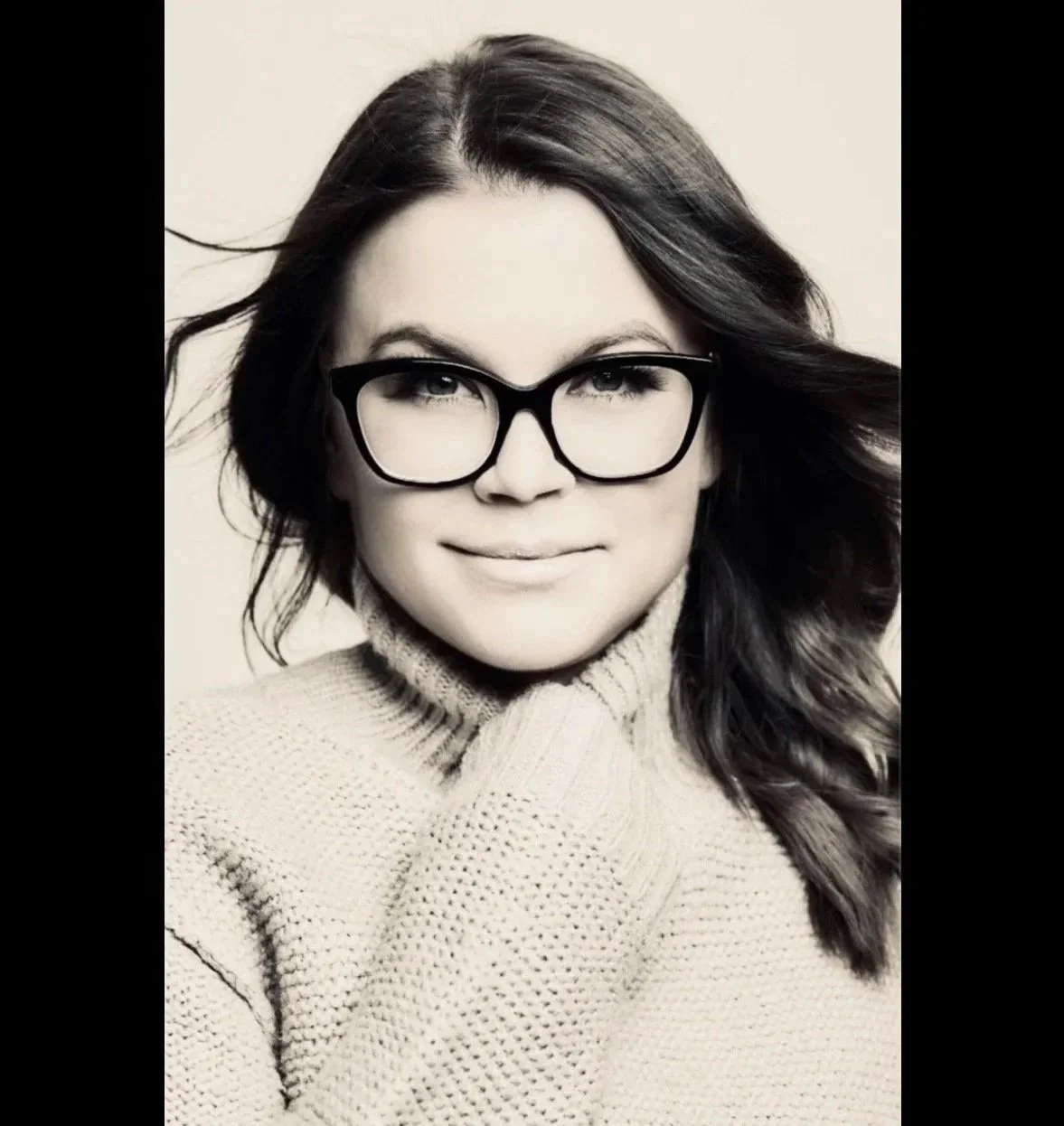 Black and white portrait of a woman with long hair, wearing glasses and a turtleneck sweater, smiling gently.