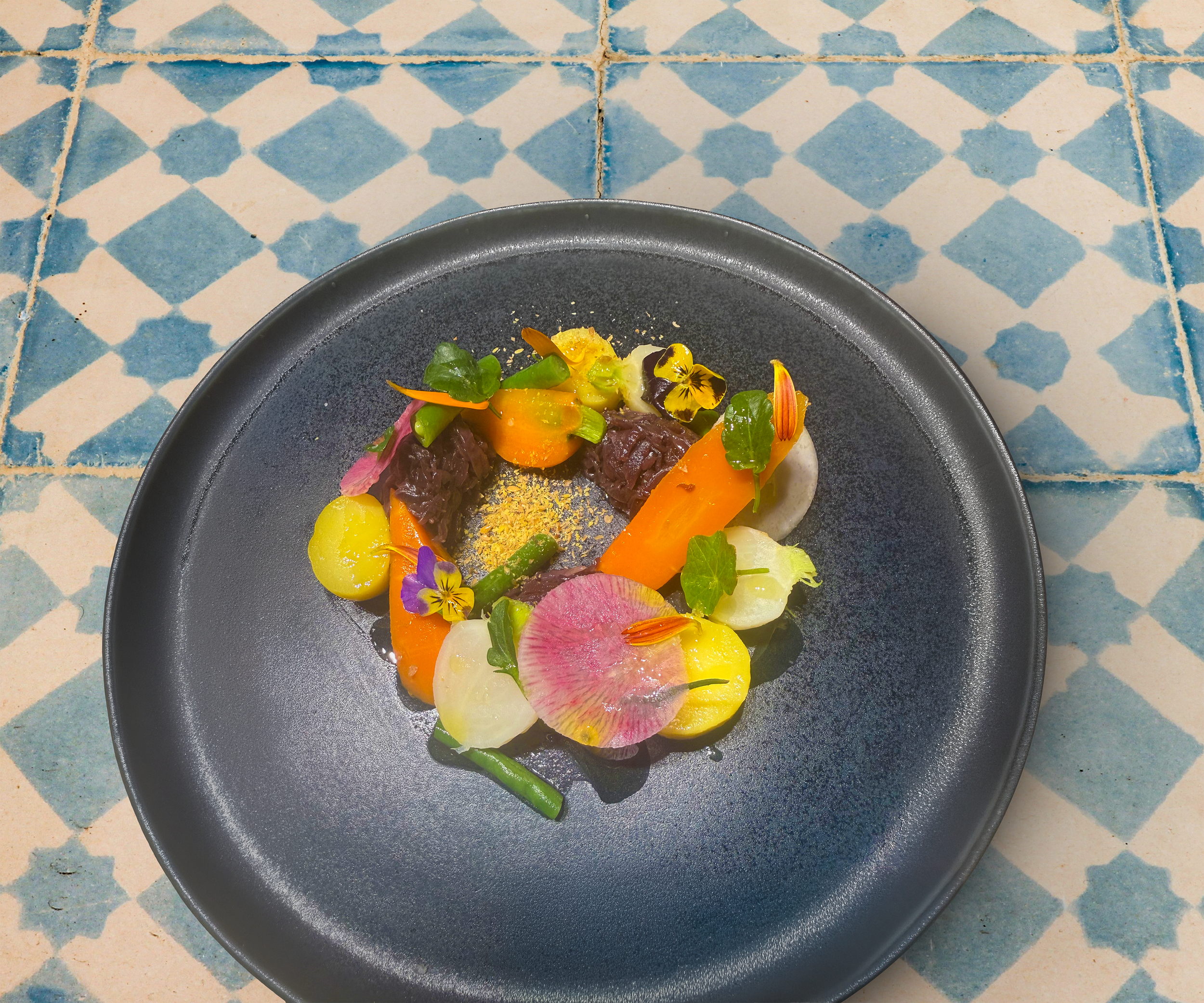 A plate with an elaborate dish garnished with various edible flowers and thinly sliced vegetables on a blue and white patterned tile table.