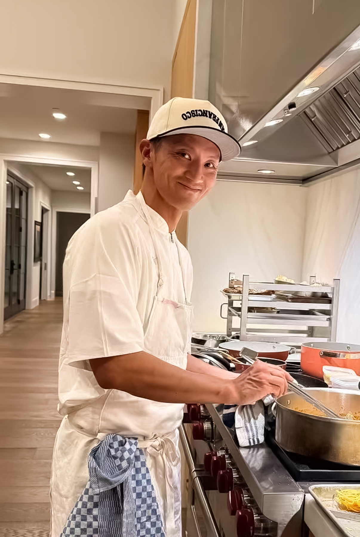 A chef in a white jacket, apron, and San Francisco baseball cap smiles over his shoulder while cooking on a professional gas range in a modern kitchen.