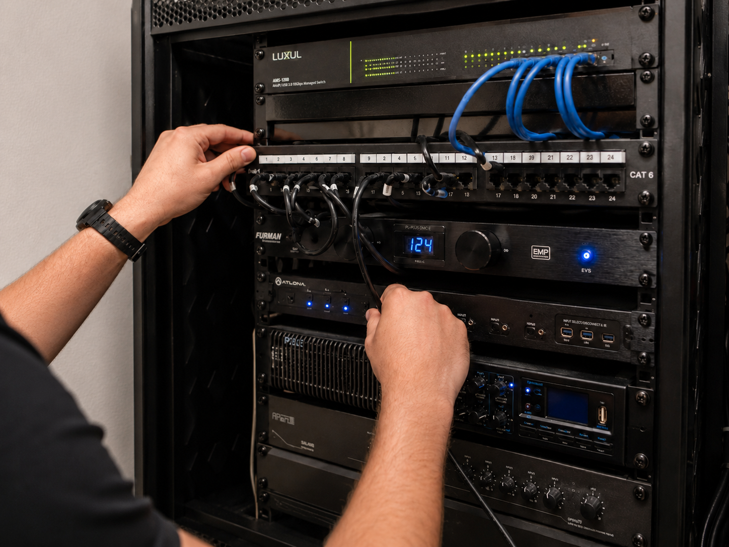 A person connecting cables to a server rack filled with networking equipment, including switches, a display with the number 124, and audio/video components.