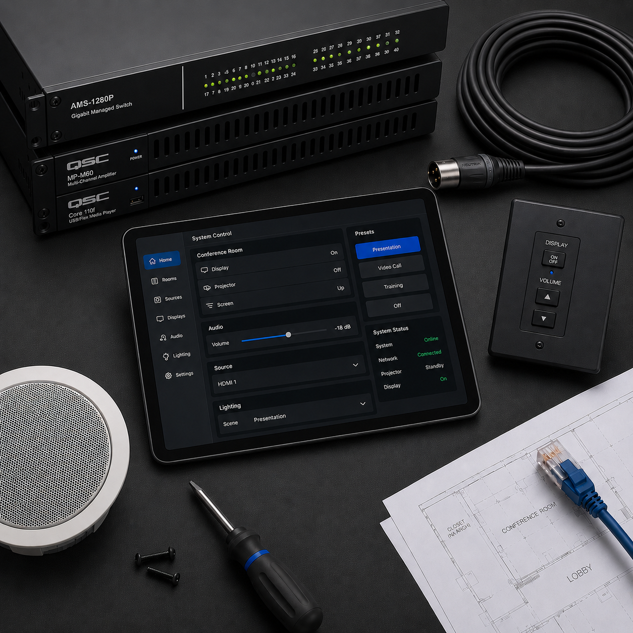 A conference room control system setup on a black desk, including a digital tablet display showing system control options, a black remote control, audio equipment, a bundle of cables, a computer networking switch, screws, a screwdriver, and architectural blueprints.
