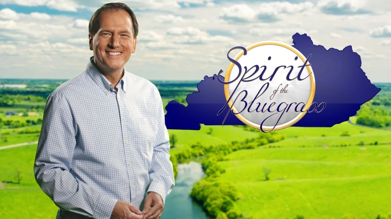 A smiling man in a light blue checkered shirt standing in front of a scenic landscape with green fields, a river, and a partly cloudy sky. To the right, there is a logo with the text 'Spirit of the Bluegrass' overlaid on a dark blue shape resembling the outline of a region.
