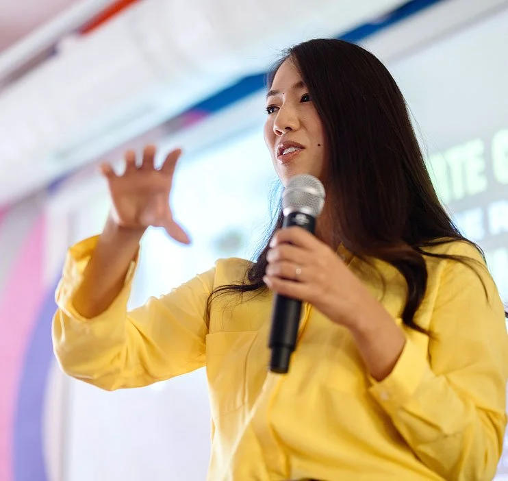 Woman in yellow shirt speaking into microphone during presentation or speech.